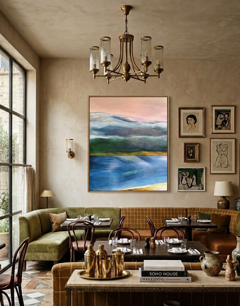 A framed landscape painting with blue, green, and pink tones hangs on a beige wall in a styled dining or lounge room with green sofas, a chandelier, and decorative vases and framed art.